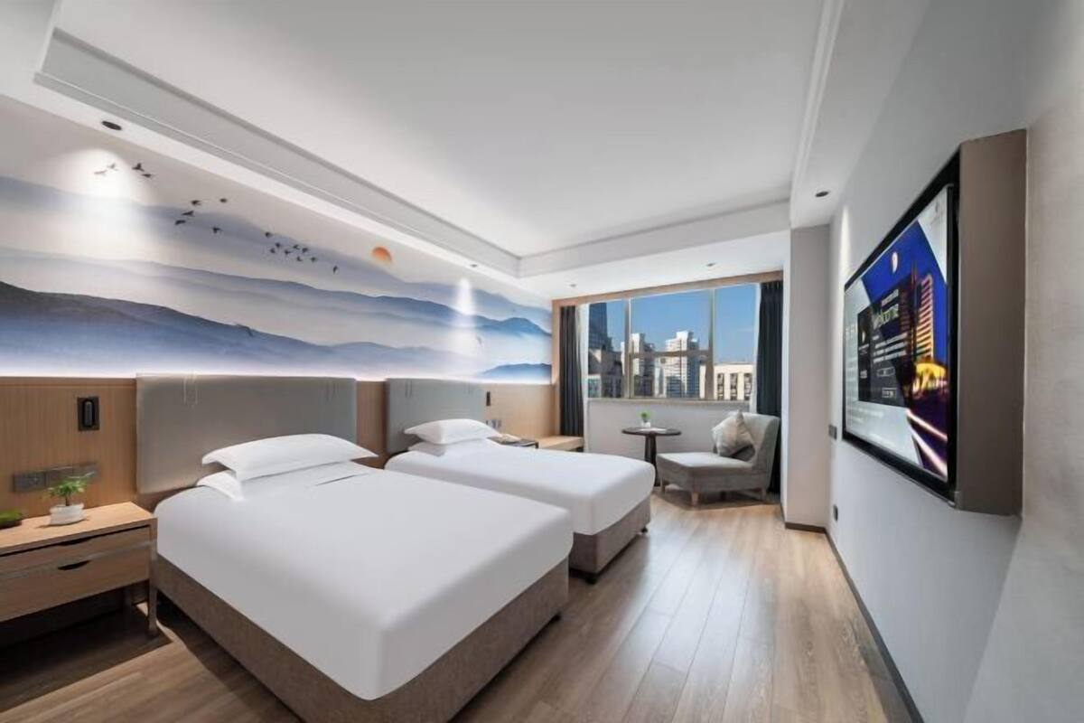 A modern hotel room with two beds, a city view, a mural, and a TV on the wall.