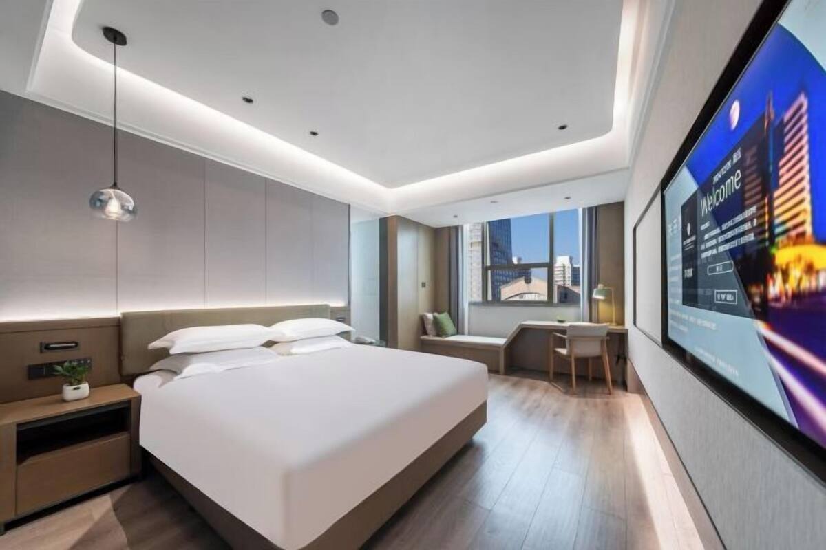 A modern hotel room with a large bed, bedside tables, a desk, and a TV screen displaying a cityscape.