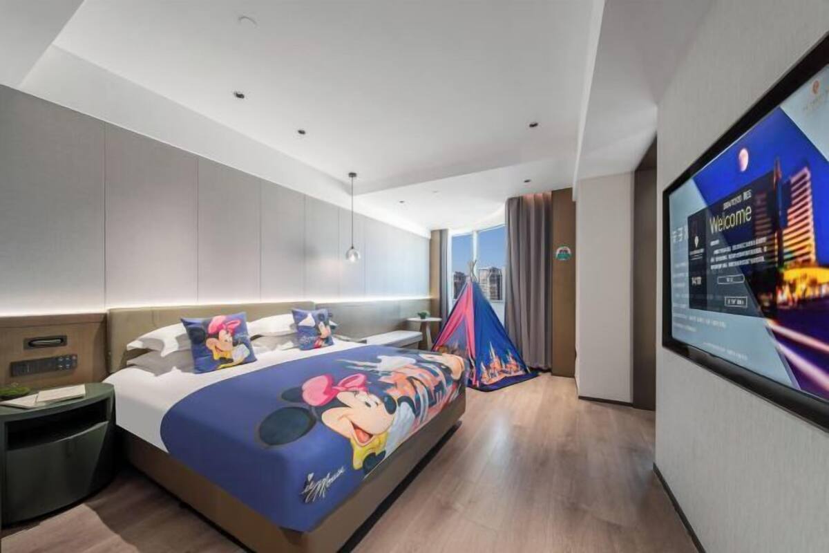 A modern hotel room with a large bed featuring a Mickey Mouse-themed bedspread, a flat-screen TV on the wall, and a tent-like structure in the corner.