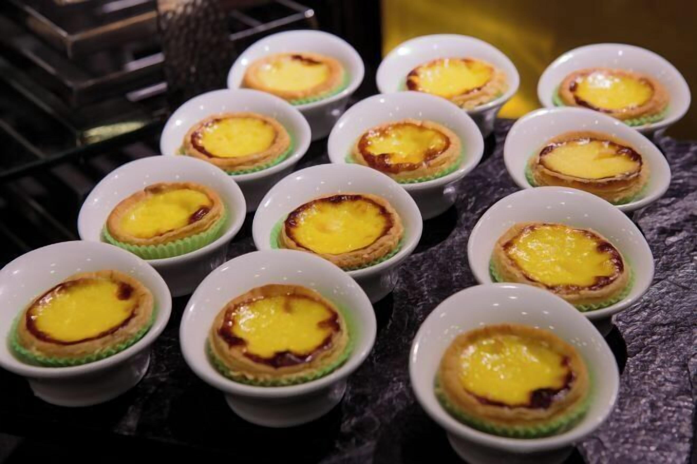 A series of small, round, white dishes with a golden-brown crust and a yellow custard filling, arranged on a dark surface.