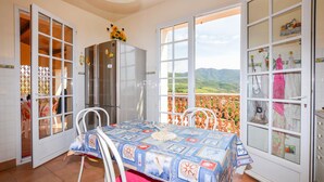 A dining area with a table covered in a patterned tablecloth, a refrigerator, and a view of a mountainous landscape through large windows. - maison vue mer panoramique (Appietto)