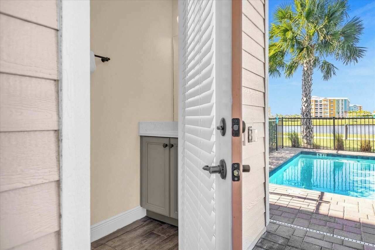 A poolside view with a door leading to a room, a small cabinet, and a view of a swimming pool and palm trees.