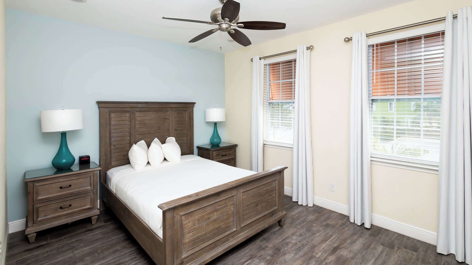 A bedroom with a wooden bed, two nightstands, a ceiling fan, and windows with blinds.