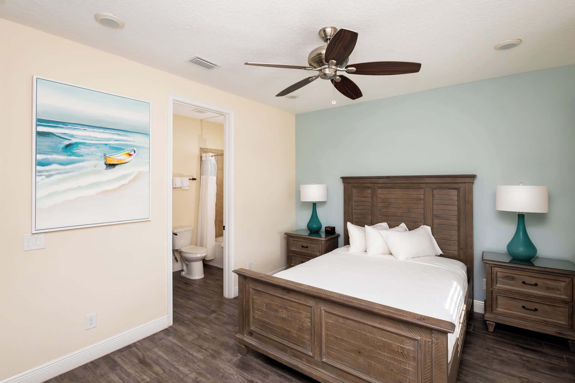 A bedroom with a wooden bed, two bedside tables with lamps, a ceiling fan, a painting of a boat on the wall, and a bathroom visible through an open door.