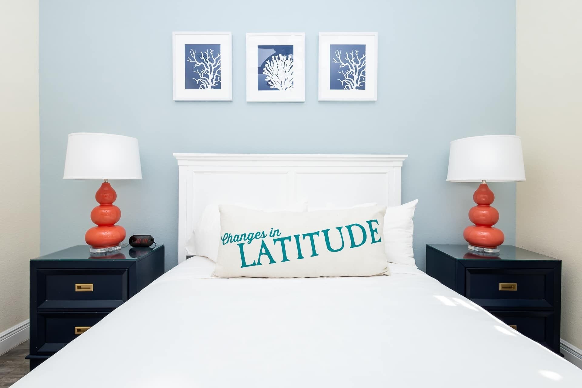 A bedroom with a white bed, two nightstands with red lamps, a white pillow with text, and three framed wall art pieces.