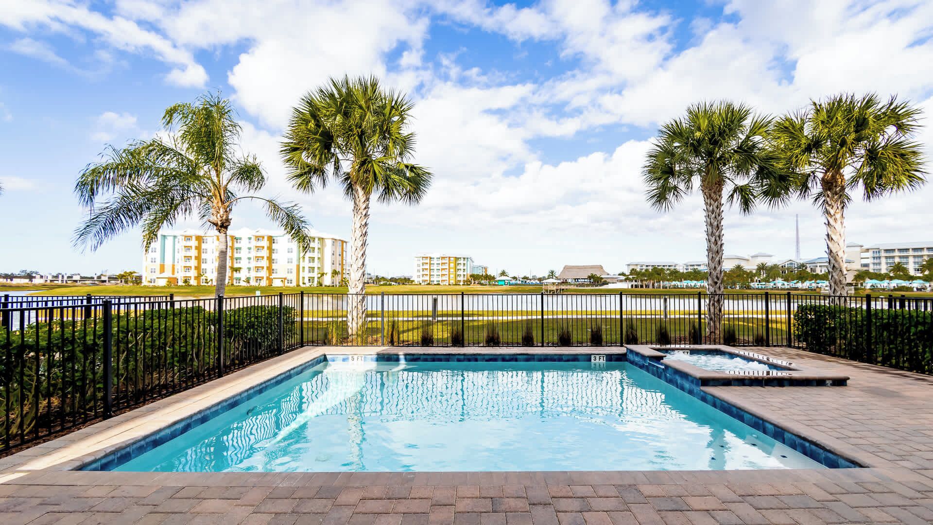 A swimming pool with a black metal fence, surrounded by palm trees and a view of buildings in the background.