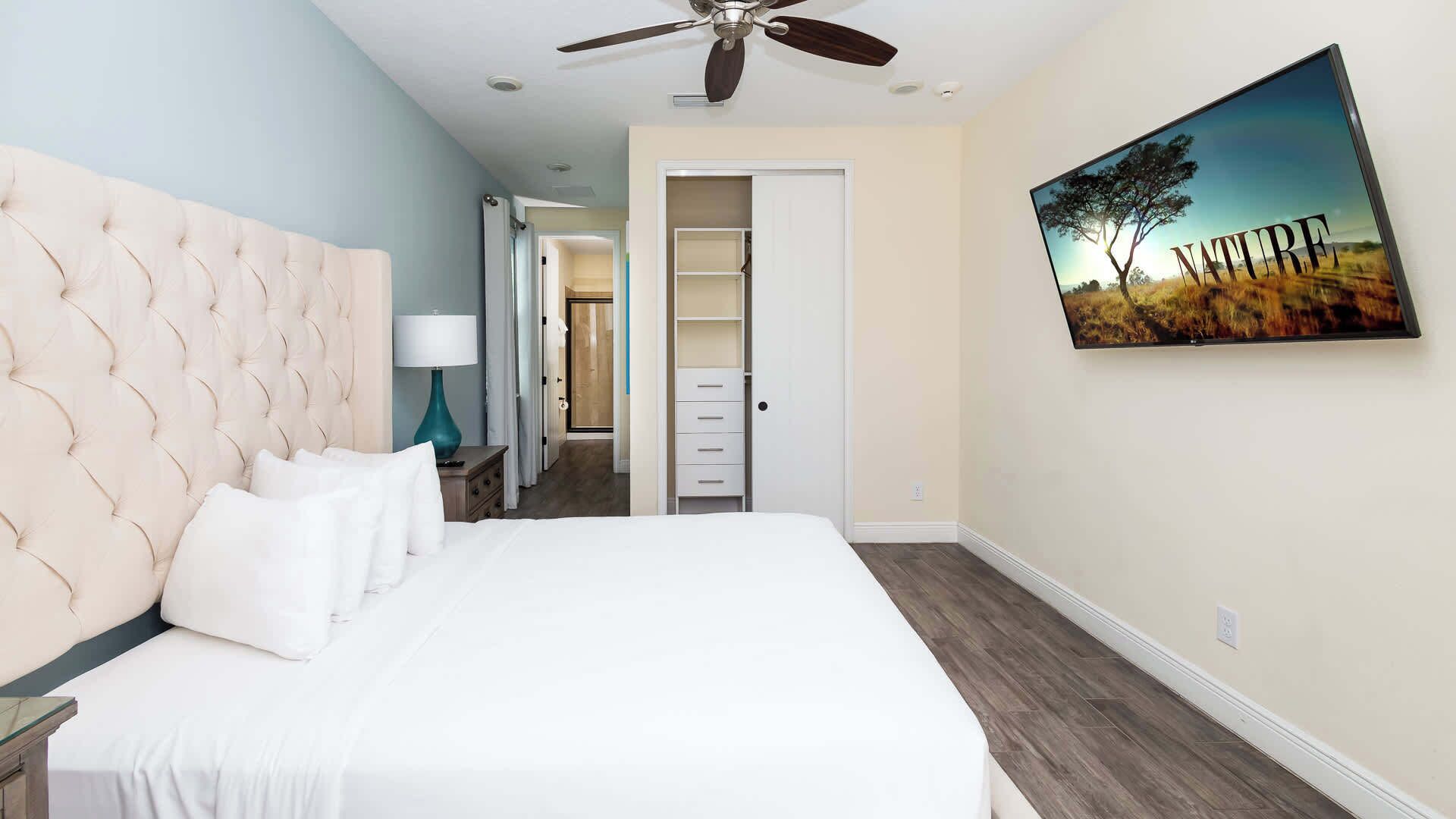A bedroom with a tufted headboard, a bed with white linens, a teal lamp, a ceiling fan, a walk-in closet, and a painting of a tree on the wall.