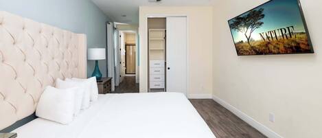 Une chambre à coucher avec une tête de lit capitonnée, un lit avec du linge de lit blanc, une lampe turquoise, un ventilateur de plafond, un dressing et un tableau représentant un arbre sur le mur.