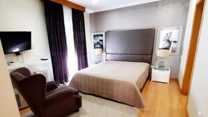 A modern hotel room with a large bed, a desk, a chair, a TV, and a small table with a lamp. - Milfontes Guest House (Odemira)