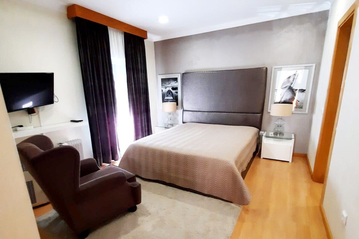A modern hotel room with a large bed, a desk, a chair, a TV, and a small table with a lamp.