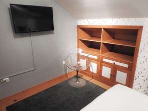 Ein Hotelzimmer mit an der Wand befestigtem Fernseher, einem Holzschrank mit mehreren Regalen, einem transparenten Stuhl und einem Bett.