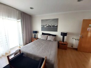 Hotelkamer met een groot bed, twee nachtkastjes met lampen, een raam met gordijnen, een radiator en een deur met nummerbord.