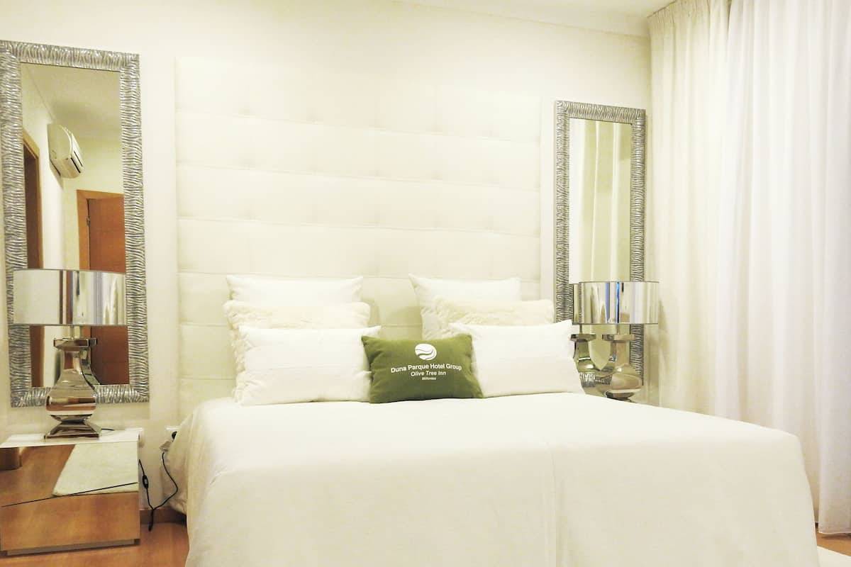 Een hotelkamer met een groot bed, twee bedlampjes, een spiegel en een kussen met het logo van het hotel.