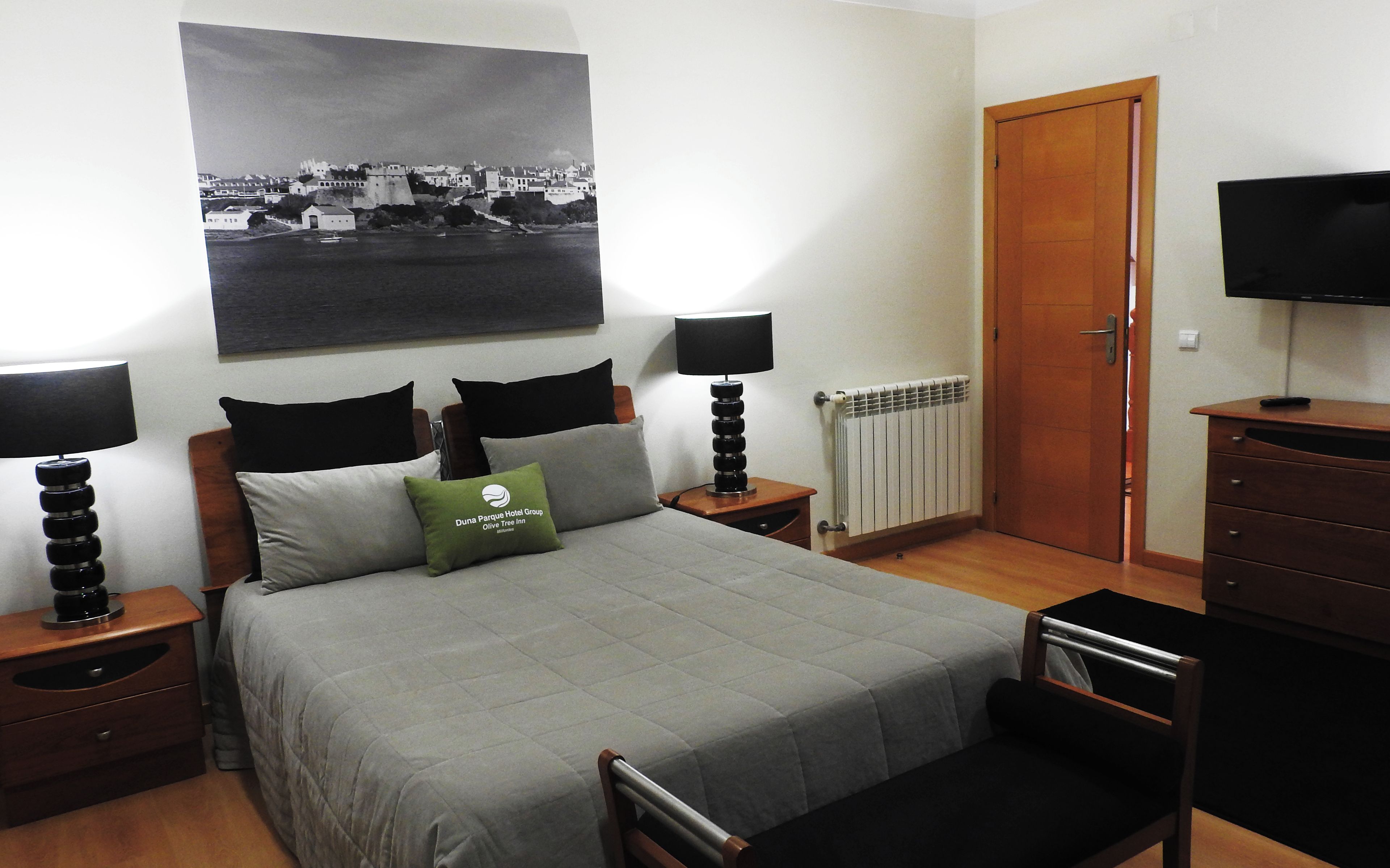 A hotel room with a bed, two bedside tables with lamps, a television, a dresser, and a large black and white cityscape painting on the wall.