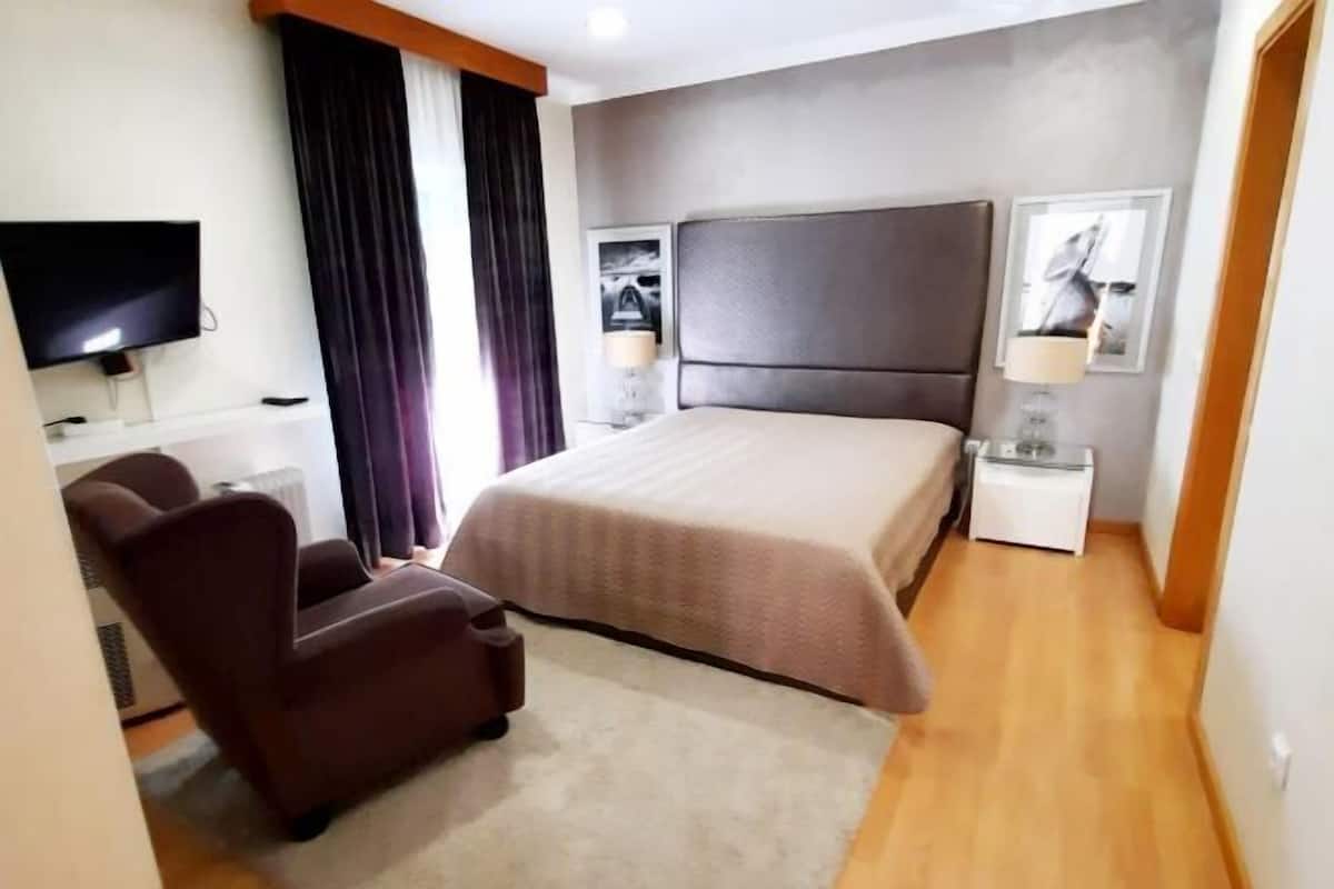 Quarto de hotel com cama, televisão, cadeira, mesa de cabeceira e janela com cortinas.