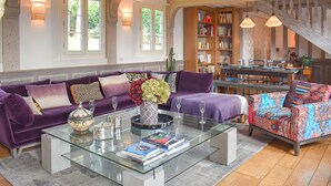Amplia sala de estar con un sofá morado, una mesa de centro de vidrio y un sillón colorido. También se observa una estantería llena de libros y una escalera.
