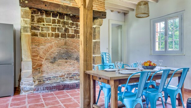 A dining area with a stone fireplace, a wooden table, blue chairs, and a window with a view of greenery.