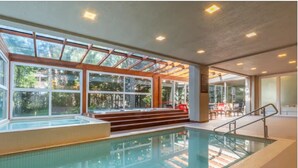 En indendørs swimmingpool med glasvægge, et trædæk og et opholdsområde med stole og et bord.