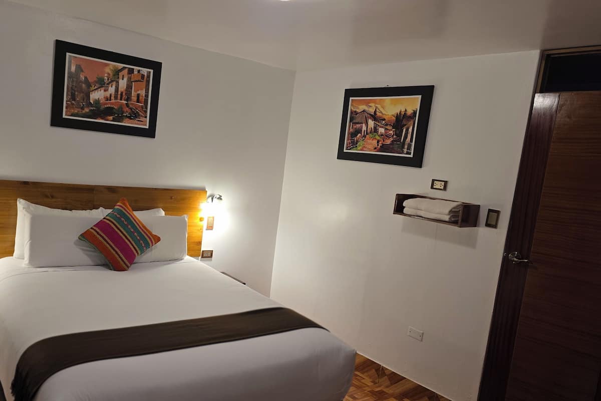 A hotel room with a wooden headboard, white bedding, a colorful pillow, two wall-mounted lights, and two framed pictures on the walls.