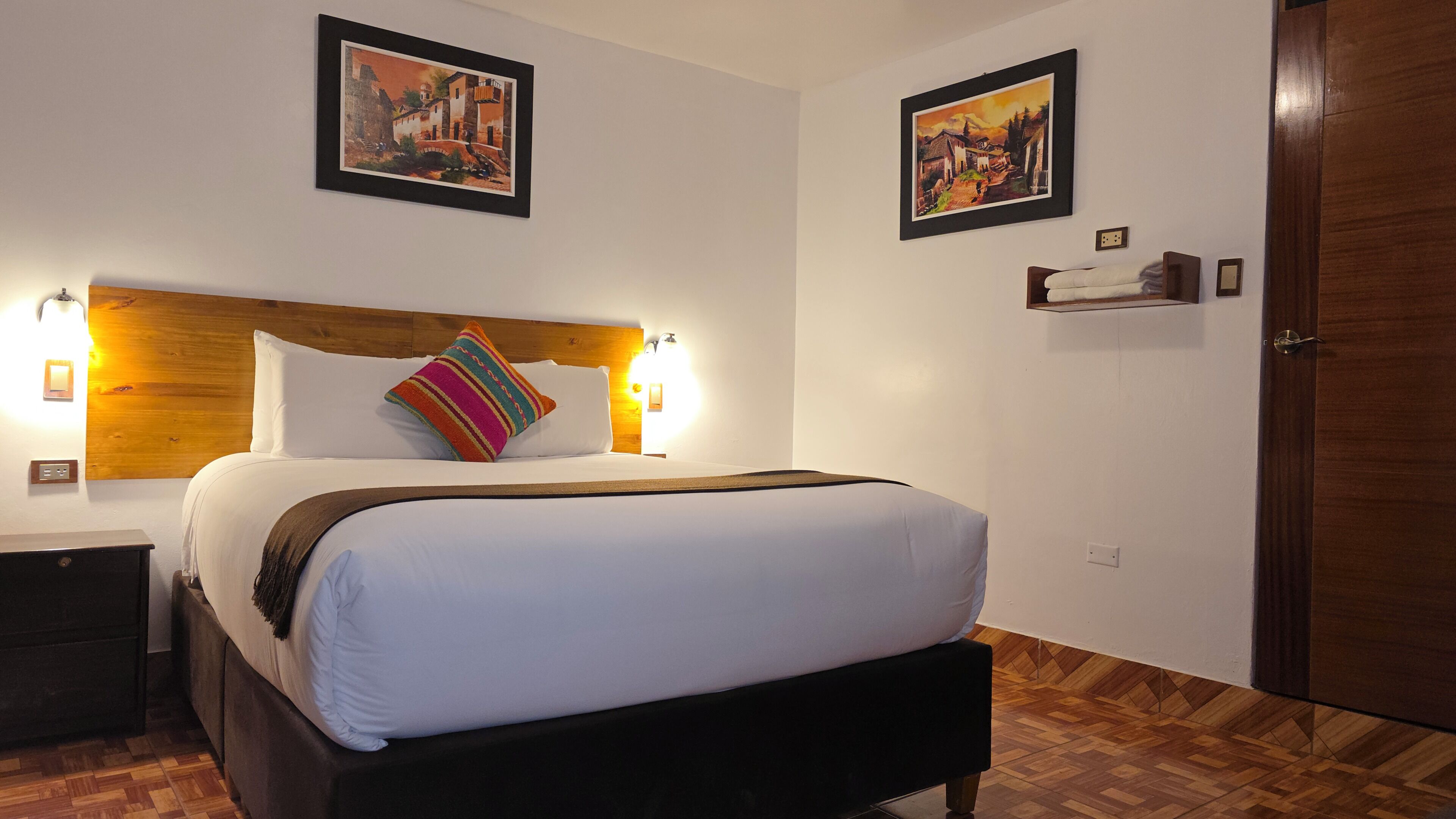 A hotel room with a large bed, wooden headboard, two wall-mounted paintings, bedside lamps, a nightstand, and a wooden floor.