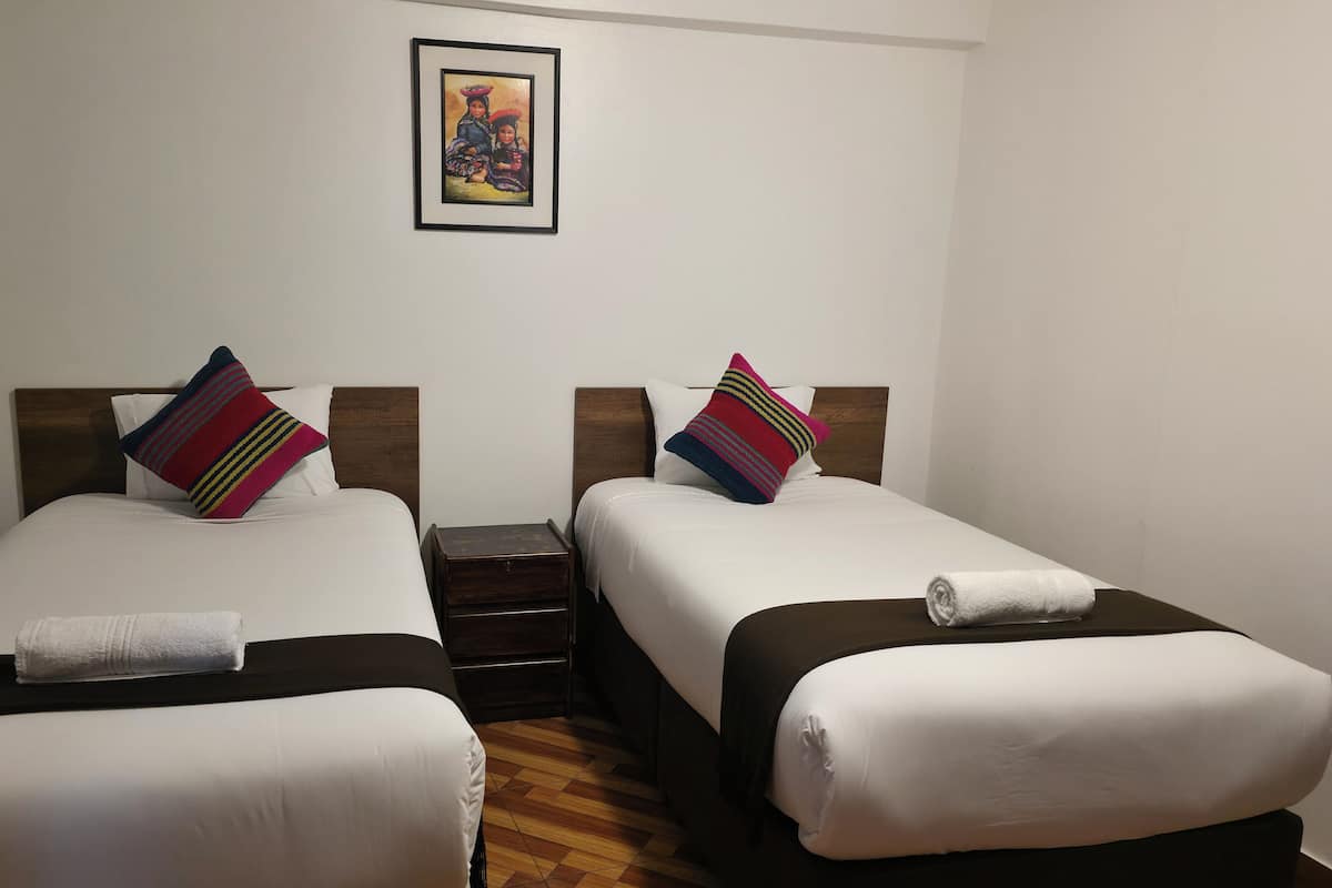 A hotel room with two single beds, each with a pillow and a rolled towel. There is a small wooden nightstand between the beds, and a framed picture hangs on the wall above.