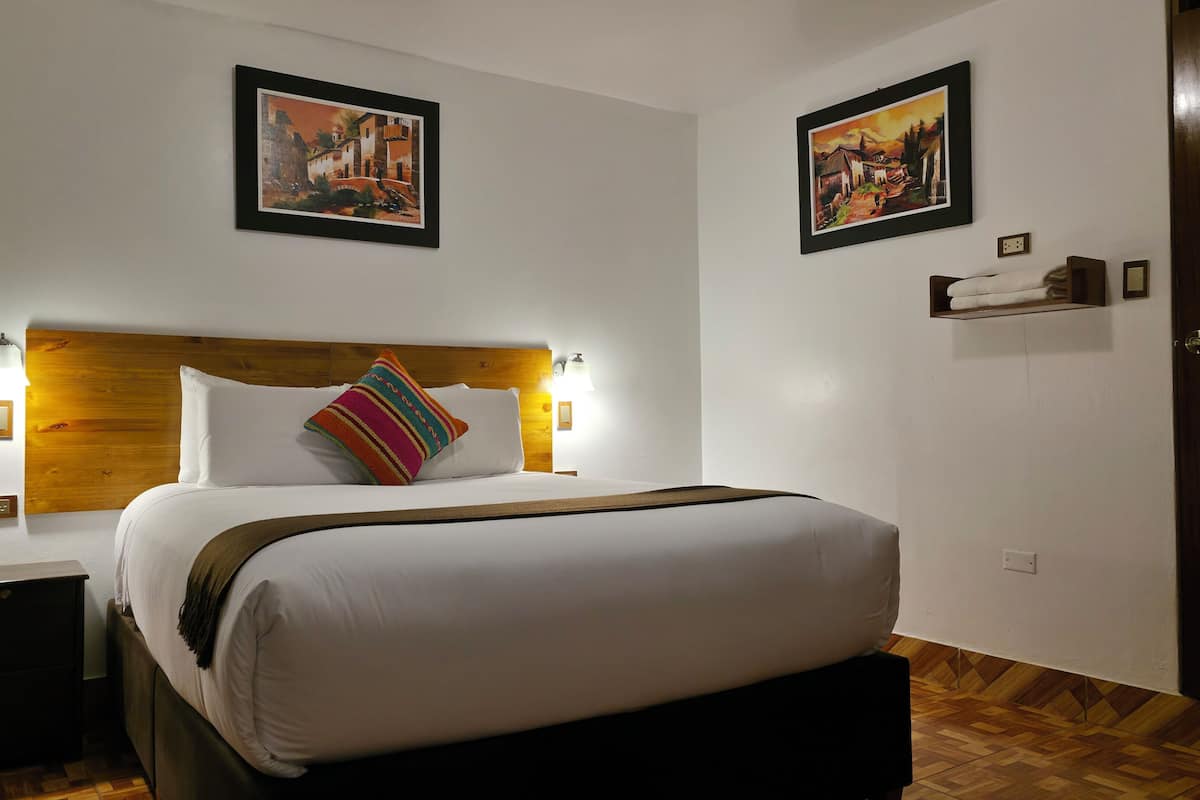 A hotel room with a wooden headboard, a bed with white linens, two wall-mounted light fixtures, and two framed pictures on the walls.