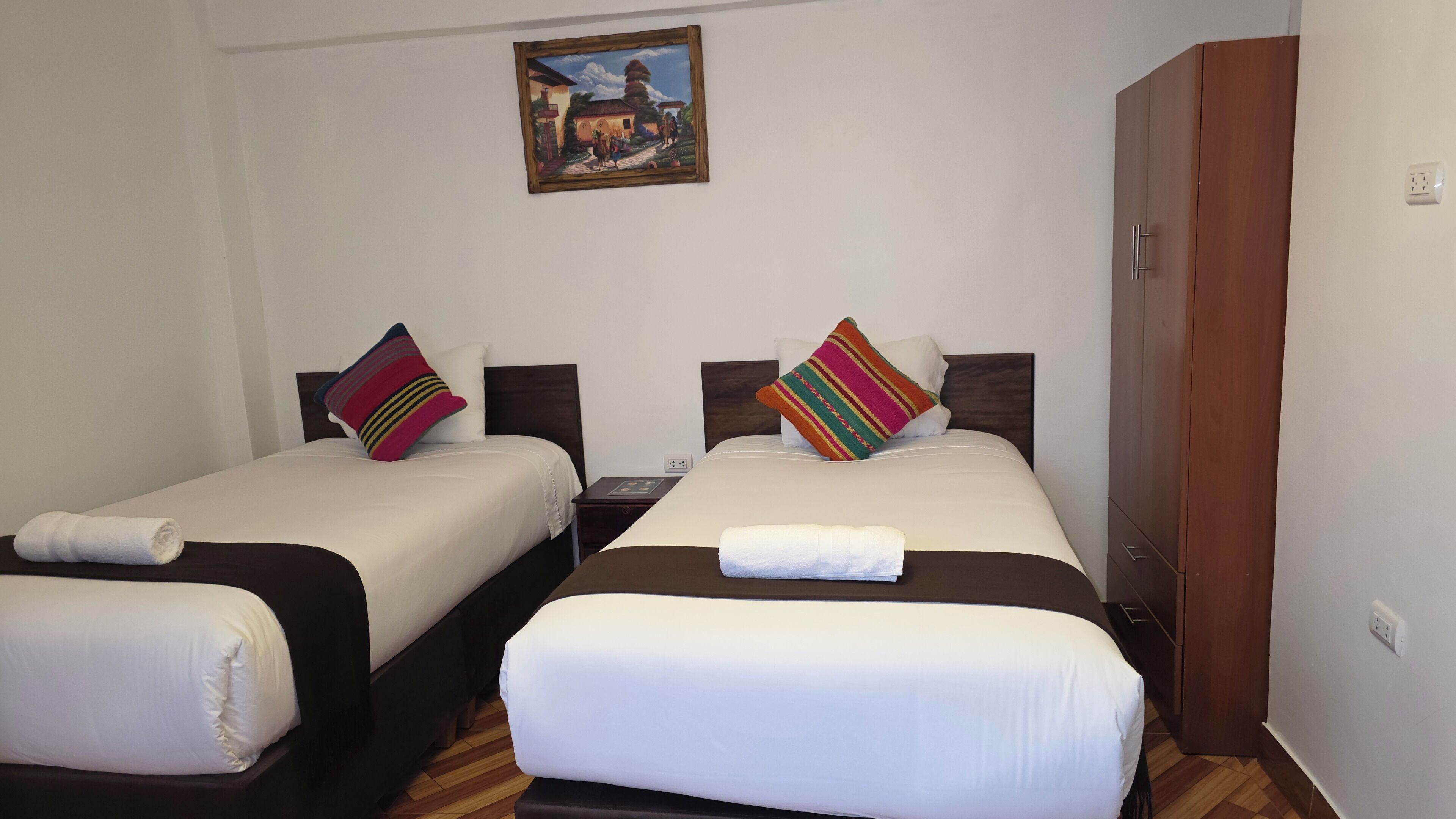 A hotel room with two beds, a wooden wardrobe, a nightstand, and a painting on the wall.