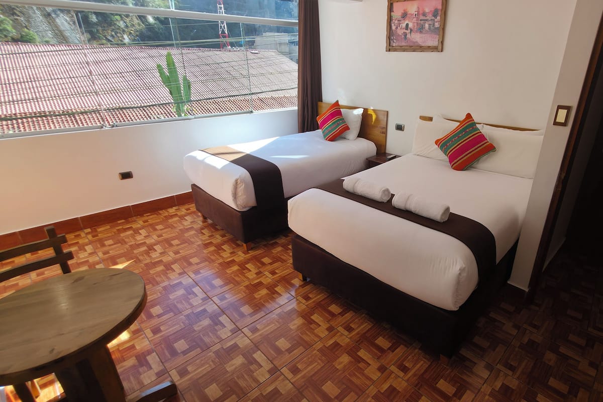 A hotel room with two beds, a small table, and a view of a red-tiled roof through a window.
