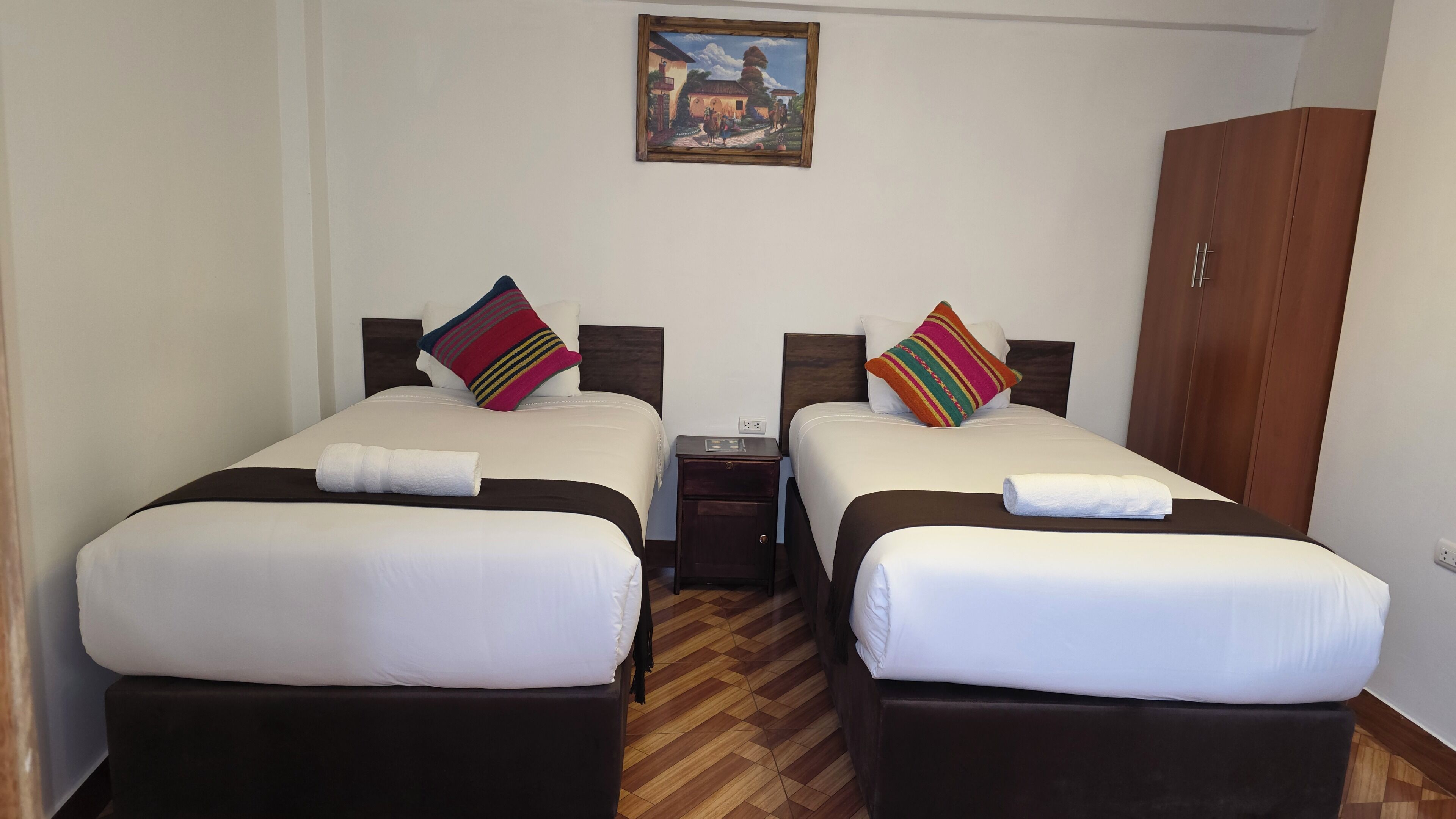 A hotel room with two beds, a wooden wardrobe, a painting on the wall, and a small bedside table.