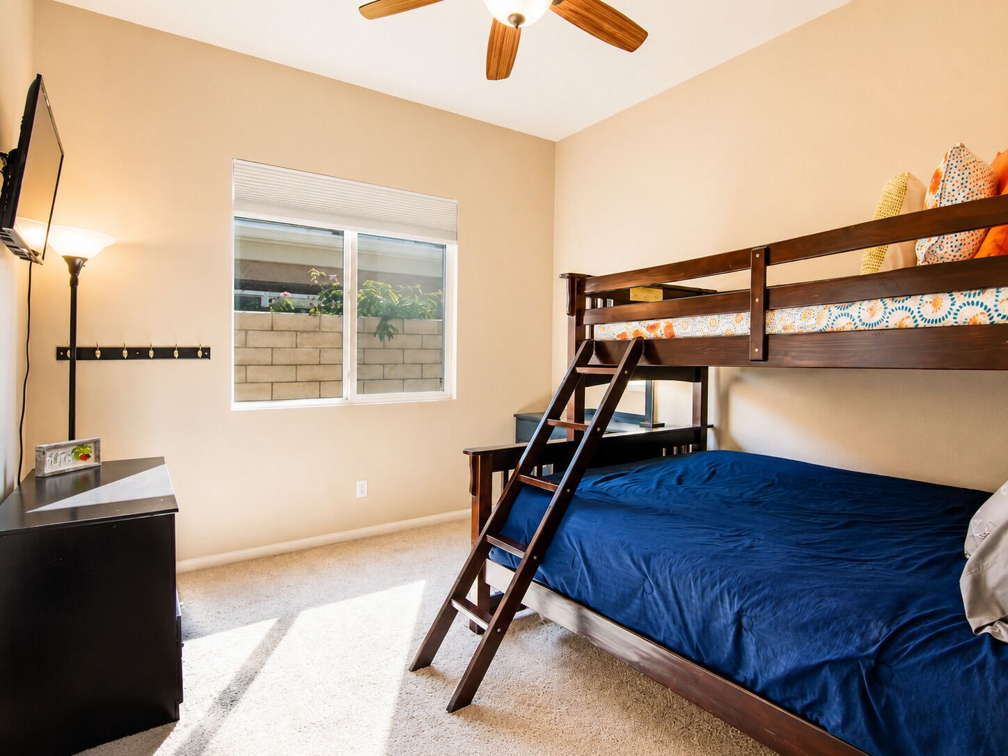 A bedroom with a bunk bed, a desk with a lamp, a window with blinds, and a ceiling fan.