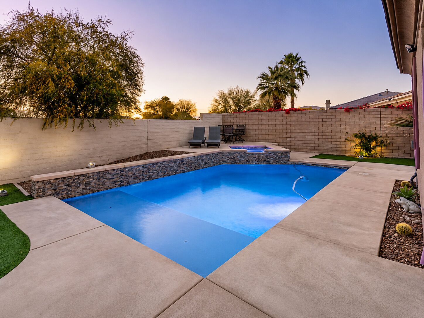 A rectangular swimming pool with a stone accent wall, surrounded by a concrete deck, a small patio area with chairs, and a landscaped garden with a palm tree.