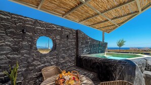 Un muro de piedra con un ventanal circular, una piscina y un área de comedor con una mesa lista para dos personas, rodeada de sillas de mimbre.