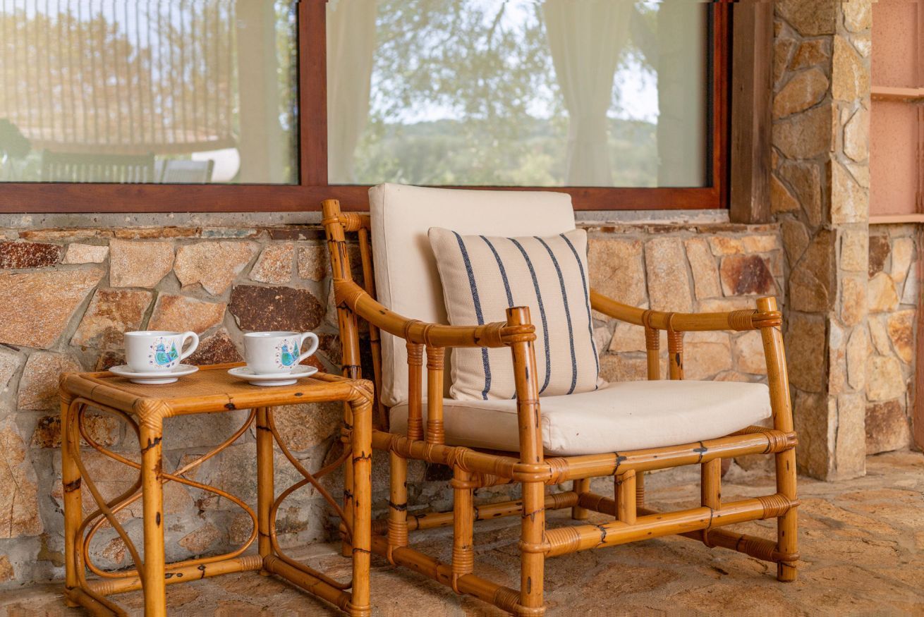 A wicker chair with a cushion and a small table with two cups, set against a stone wall with a window above.