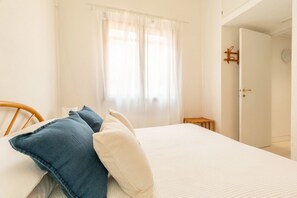 A neatly made bed with white and blue pillows, a wooden headboard, and a window with sheer curtains. - BLUE BAY HOME just few steps from the bay of Porto Luna (Villasimius)