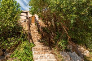Un escalier en pierre mène à un bâtiment dont le toit est recouvert de tuiles, et qui est entouré de verdure.