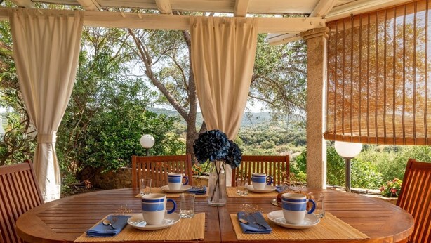 Mesa de madeira redonda posta para quatro, com xícaras azuis e brancas, um vaso com flores azuis e uma pérgola de madeira com cortinas bege.