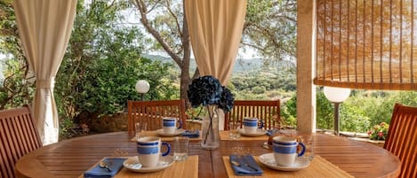 Un tavolo di legno rotondo apparecchiato per quattro, con tazze bianche e azzurre, un vaso con fiori blu e un pergolato di legno con tende beige.