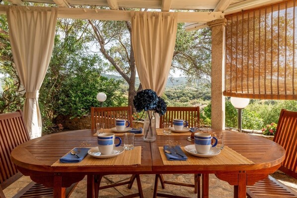 A round wooden table set for four with blue and white cups, a vase with blue flowers, and a wooden pergola with beige curtains.