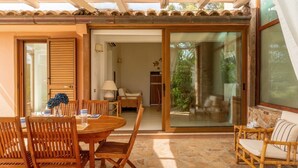 Um pátio ensolarado com mesa e cadeiras de madeira, uma porta de correr de vidro que dá para um jardim e piso de pedra.