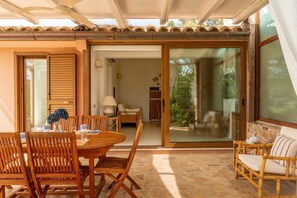 A sunlit patio with a wooden table and chairs, a glass sliding door leading to a garden, and a stone floor.