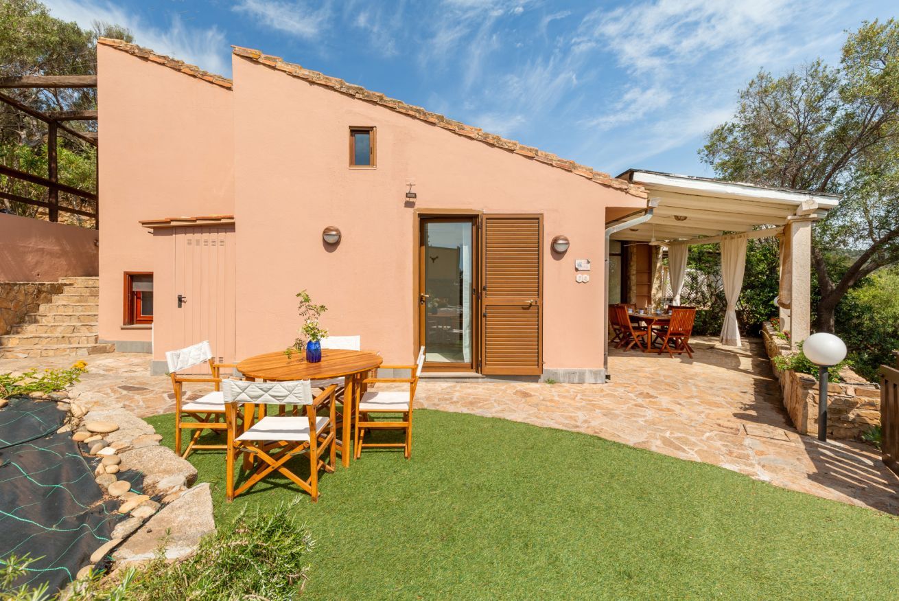 Une maison à deux étages dotée d’un patio comprenant une table et des chaises en bois, un espace salon couvert et un chemin en pierre menant à l’entrée.