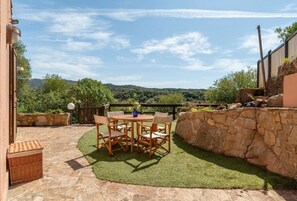 A patio with a stone table and chairs, surrounded by a stone wall and a grassy area with a view of hills.