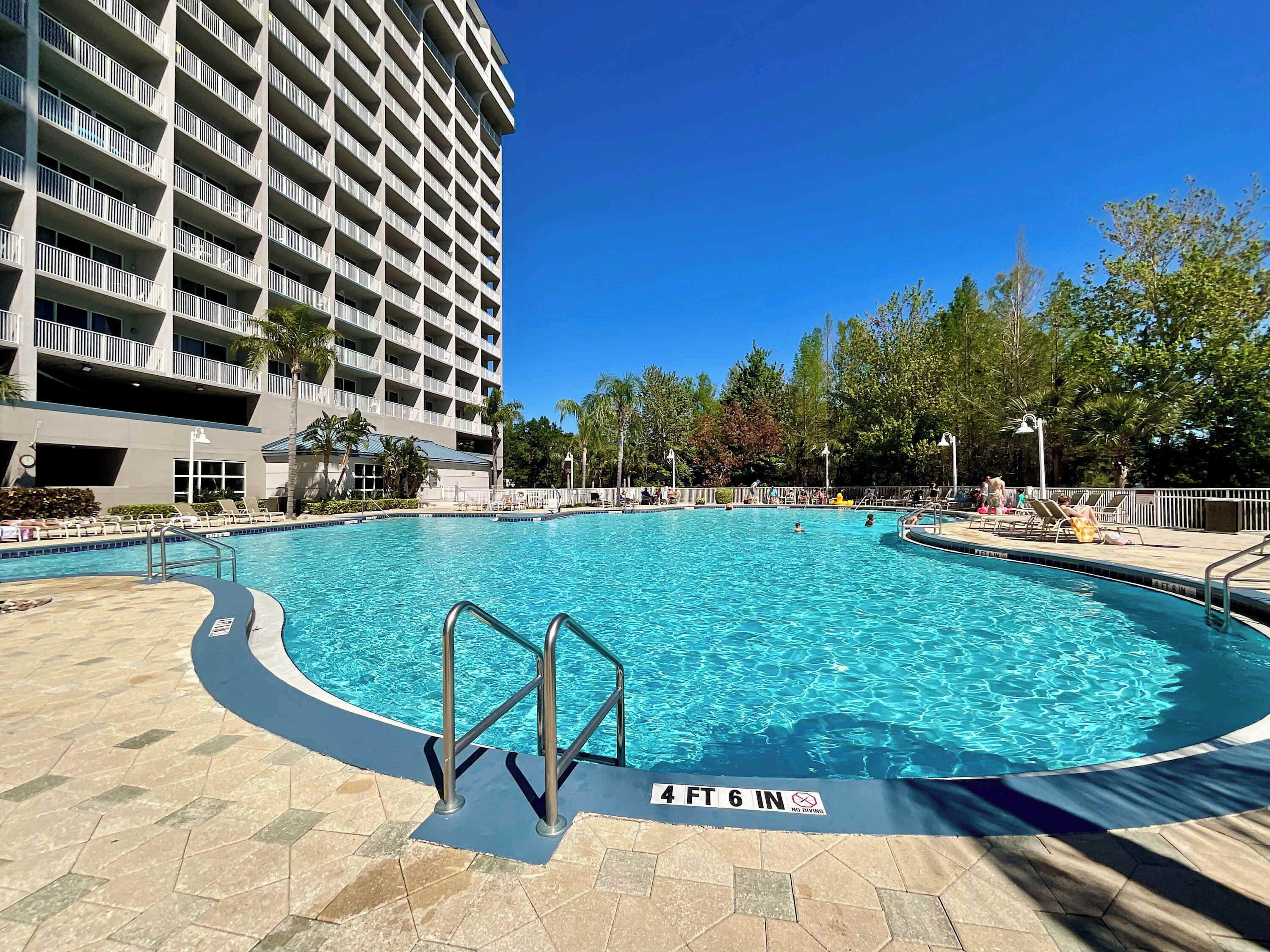 A swimming pool with a 4 ft 6 in depth sign, surrounded by lounge chairs and a multi-story building.