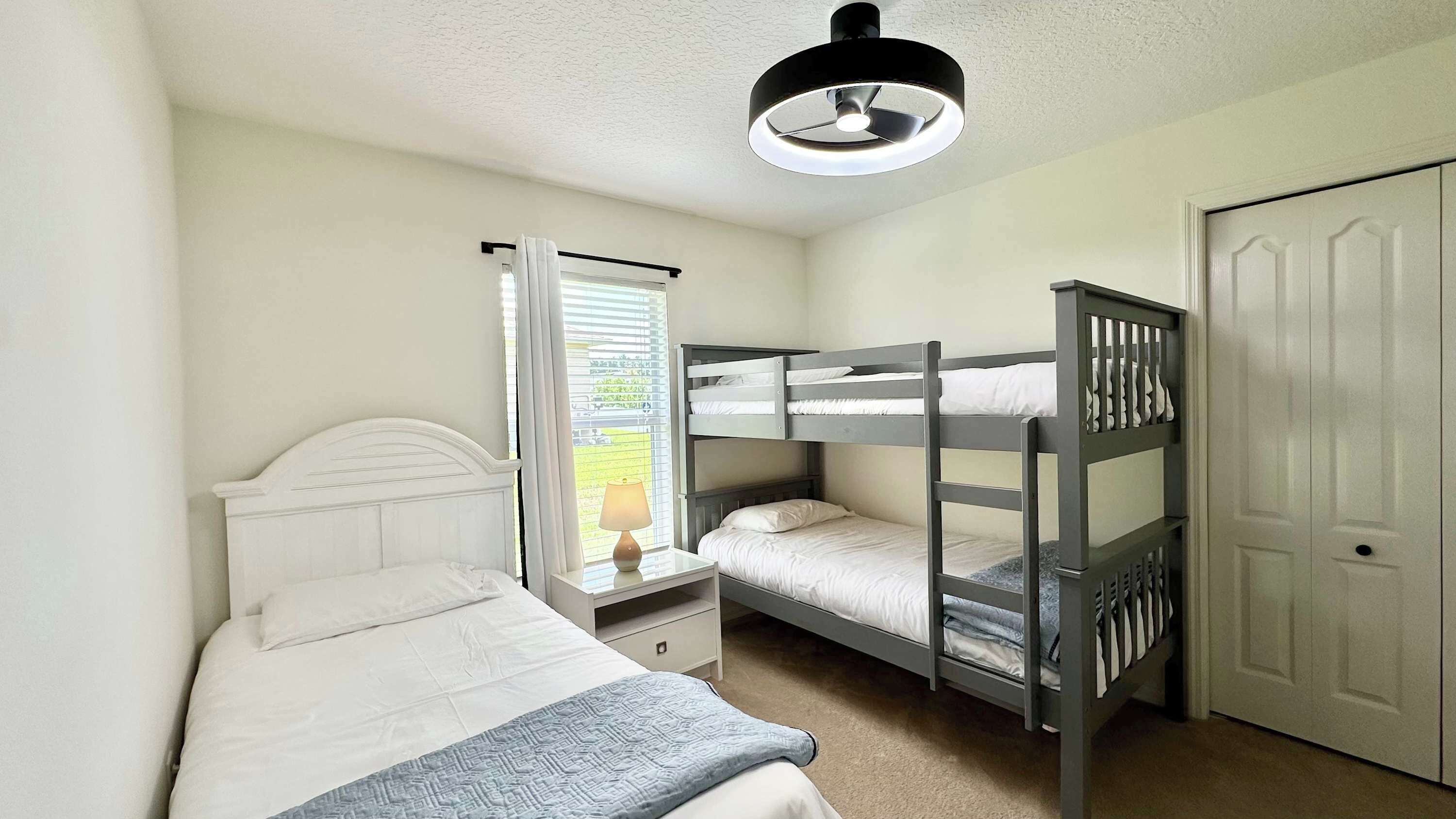 A bedroom with a bunk bed, a single bed, a nightstand, a lamp, a window with curtains, and a door.