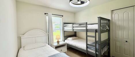 A bedroom with a bunk bed, a single bed, a nightstand, a lamp, a window with curtains, and a door.