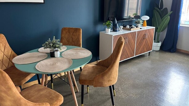 Een eetgedeelte met een blauwgroene tafel, vier stoelen, een wit kastje en een televisie aan de muur.