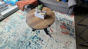 Een moderne woonkamer met een grijze bank, kleurrijke kussens, een ronde salontafel met boeken en een plant, en abstracte kunst aan de muur.