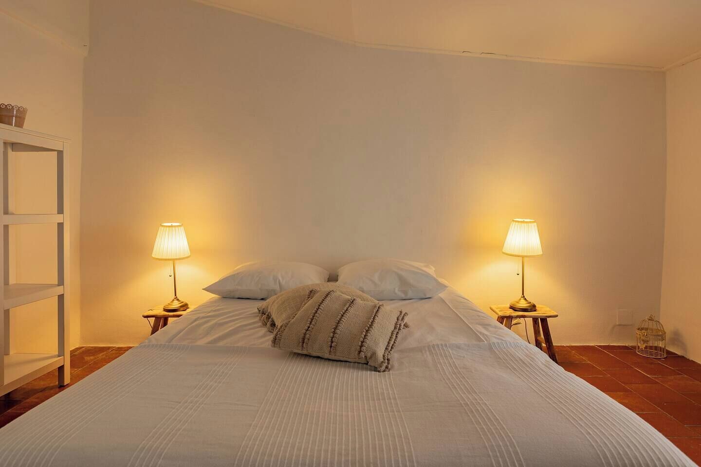 Una cama bien hecha, con dos lámparas de noche, una mesita de madera y una estantería blanca contra una pared lisa.