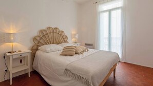 A bedroom with a large bed, two bedside tables with lamps, a window with sheer curtains, and a wooden floor. - The discreet 18th century (Sérignan)