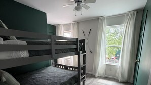Una habitación con literas, ventilador de techo, ventana con cortinas y una puerta.